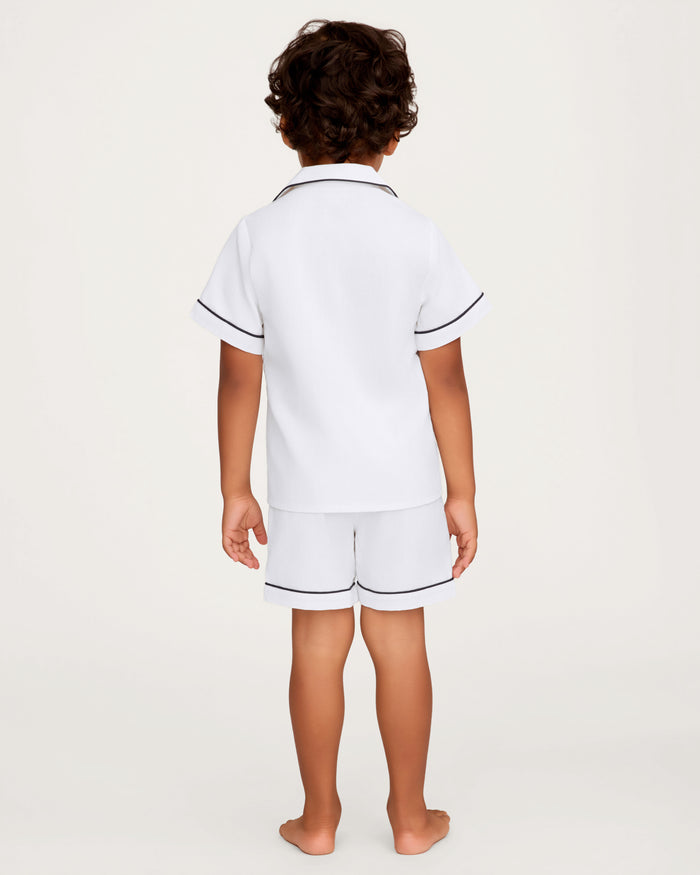 A young child with curly hair stands barefoot, facing away, wearing the Kid’s Twill Pajama Short Set in White with Yacht Club Embroidery, featuring dark piping on the edges, against a plain light background.
