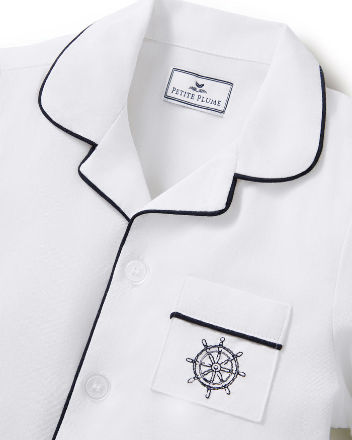 Kid’s Twill Pajama Short Set in White with Yacht Club Embroidery features a white pajama top with black piping, buttons, and a front pocket embroidered with a navy ship wheel. The label inside reads Petite Plume.