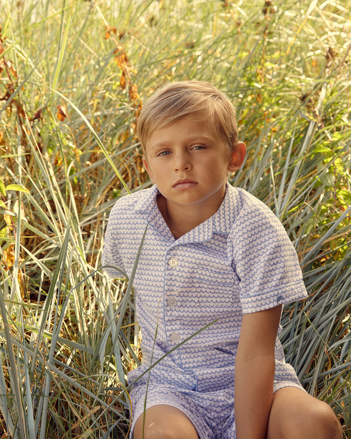 A young boy with short blond hair sits in tall grass, wearing Petite Plumes Kids Twill Pajama Short Set in La Mer. Made from the finest cotton, he gazes at the camera neutrally. The backdrop is vibrant with green and yellow grasses.