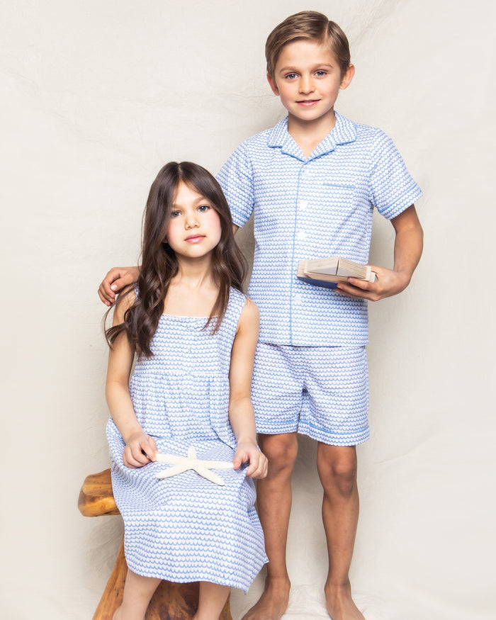A girl in a blue dress holds a starfish, while the boy, dressed in a Kids Twill Pajama Short Set in La Mer by Petite Plume, cradles a small wrapped gift. They stand together on a wooden stool against a plain background, capturing the charm of simple childhood moments.