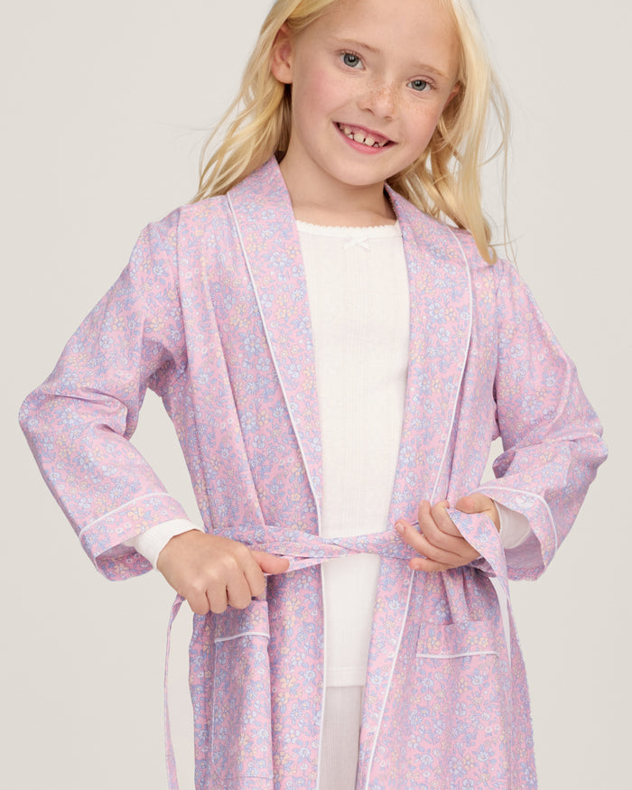 A young girl with blonde hair smiles as she ties the belt on her Kid's Twill Robe in Fleur Blush over a white nightgown.