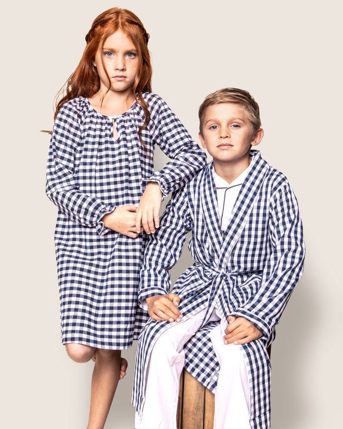 Two children wear Petite Plumes matching navy gingham sleepwear. The girl has a long nightgown, and the boy wears pajamas topped with the Kids Twill Robe in Navy Gingham. They stand calmly against a plain backdrop, highlighting neutral expressions and childrens sleepwear.