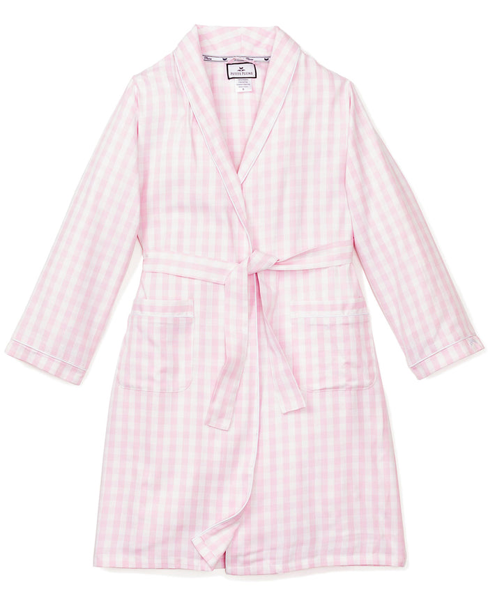 A close-up of Petite Plumes Kids Twill Robe in Pink Gingham shows its soft, textured fabric, reminiscent of classic childrens sleepwear. The robe features a folded cuff with stitched letters and a tied belt across the light pink and white gingham pattern.