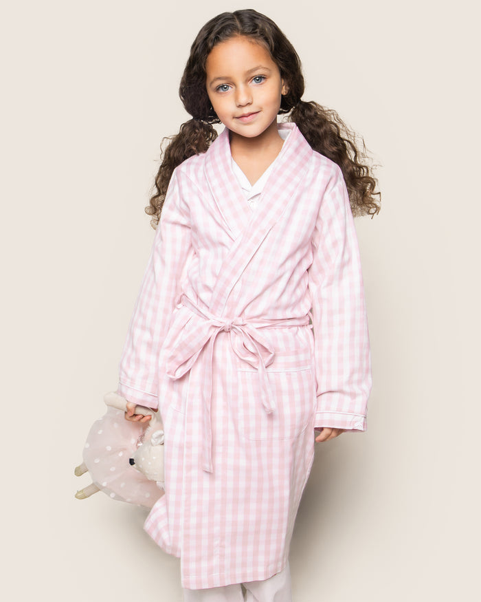 A close-up of Petite Plumes Kids Twill Robe in Pink Gingham shows its soft, textured fabric, reminiscent of classic childrens sleepwear. The robe features a folded cuff with stitched letters and a tied belt across the light pink and white gingham pattern.
