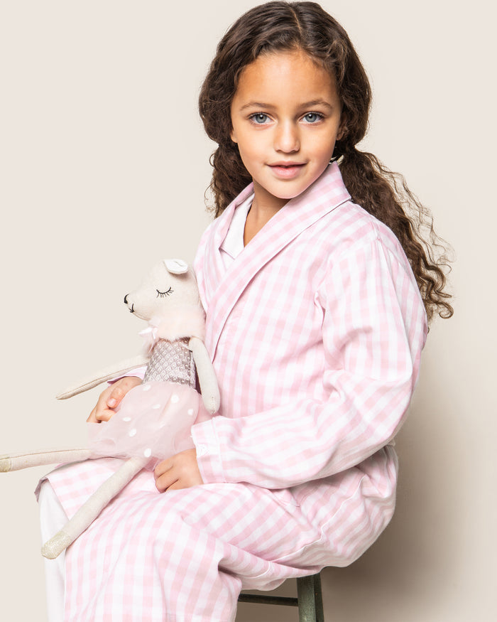 A close-up of Petite Plumes Kids Twill Robe in Pink Gingham shows its soft, textured fabric, reminiscent of classic childrens sleepwear. The robe features a folded cuff with stitched letters and a tied belt across the light pink and white gingham pattern.