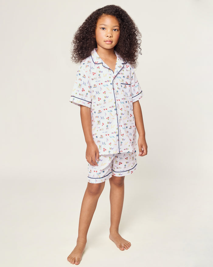 The Kid's Twill Short Set in Literary Lessons features a white base with colorful school-themed prints—pencils, books, and erasers—plus navy piping on the collar, pocket, sleeves, and shorts.
