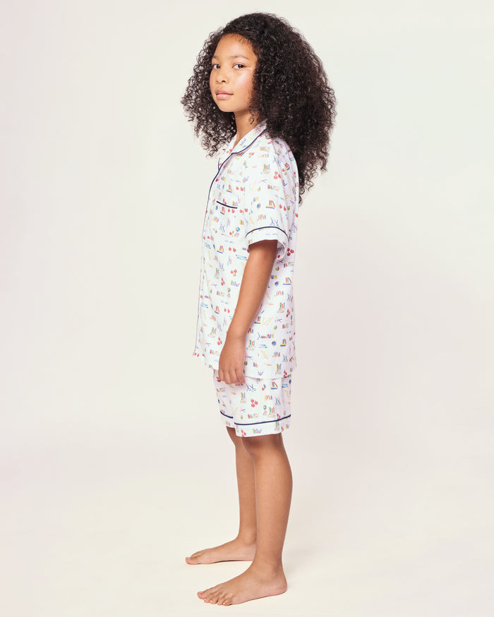 The Kid's Twill Short Set in Literary Lessons features a white base with colorful school-themed prints—pencils, books, and erasers—plus navy piping on the collar, pocket, sleeves, and shorts.
