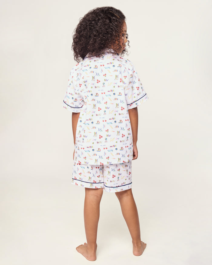 The Kid's Twill Short Set in Literary Lessons features a white base with colorful school-themed prints—pencils, books, and erasers—plus navy piping on the collar, pocket, sleeves, and shorts.