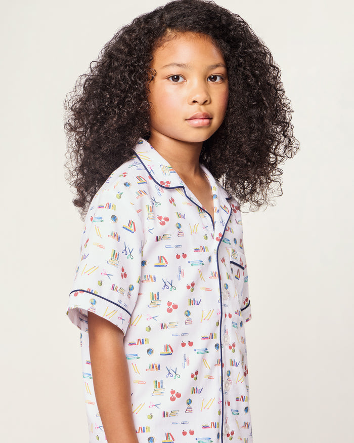 The Kid's Twill Short Set in Literary Lessons features a white base with colorful school-themed prints—pencils, books, and erasers—plus navy piping on the collar, pocket, sleeves, and shorts.