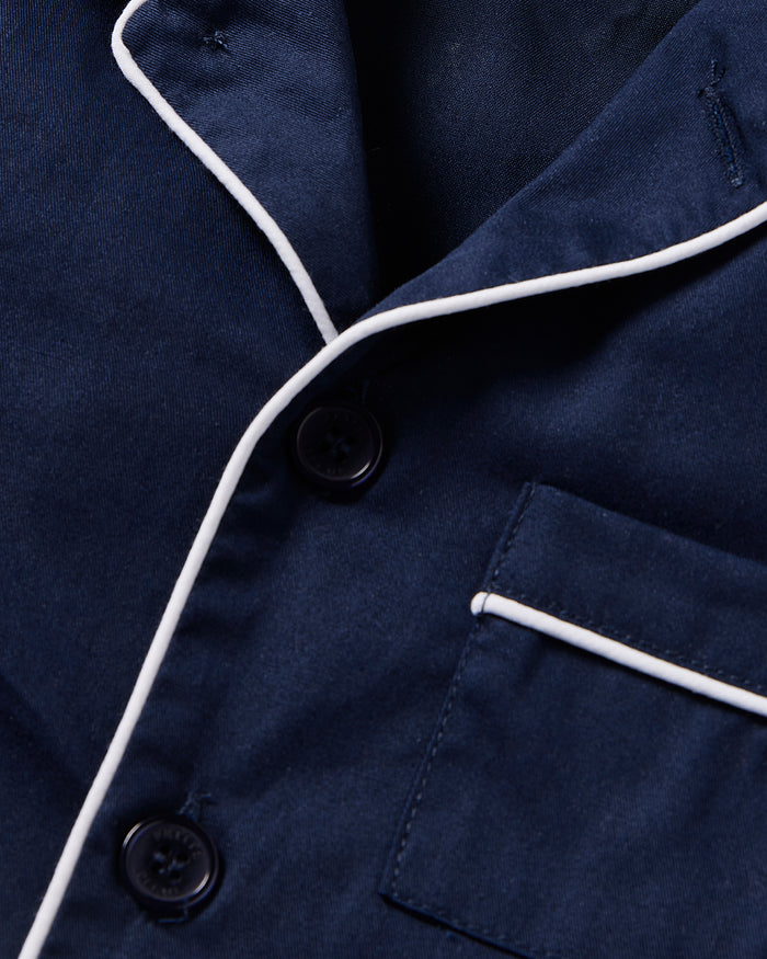 The close-up features Petite Plumes Kids Twill Short Sleeve Short Set in Navy, showcasing navy fabric with white piping, two black buttons, and a pocket accented by matching white trim, creating an elegantly refined sleepwear piece.