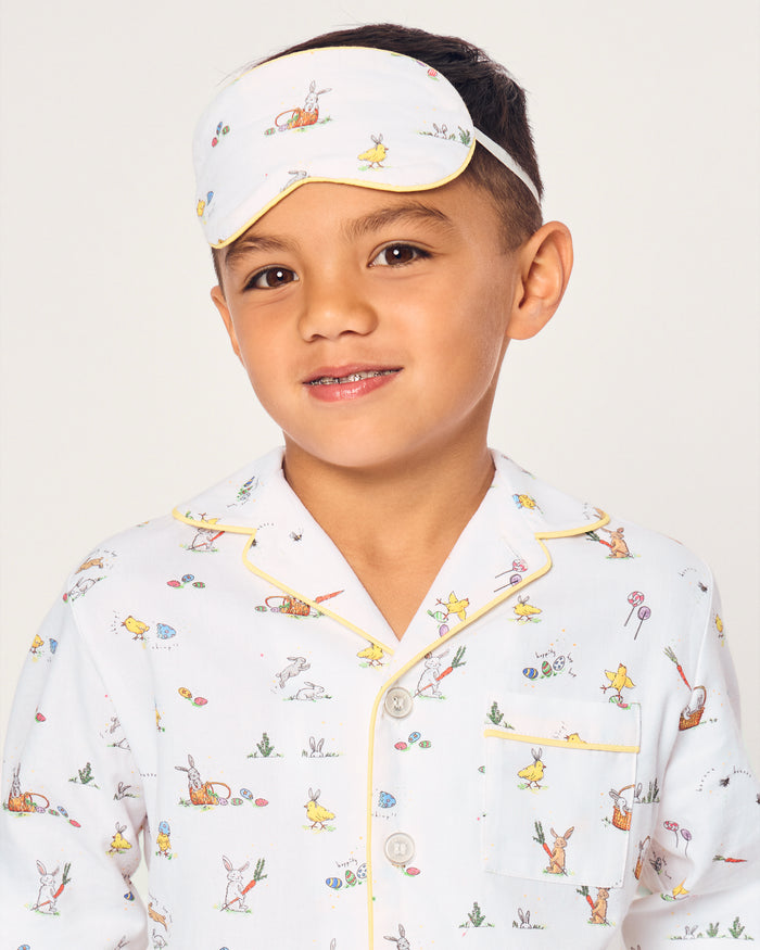 A young boy wears a Kid's Twill Sleep Mask in Easter Frolic, featuring colorful animal and nature prints, along with matching white pajamas. He smiles softly against a plain, light background.