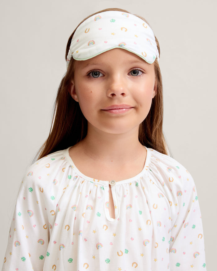 A young girl with long brown hair wears the Kid's Twill Sleep Mask in Good Luck Charms and a matching pajama top featuring rainbows, horseshoes, and clovers, standing against a neutral background.