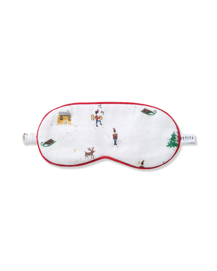 The Kid's Twill Sleep Mask in Holiday Town is a white mask with red trim, decorated with winter scenes of people, a reindeer, a house, a sled, and snowy trees. A small white tag is attached on the right side.