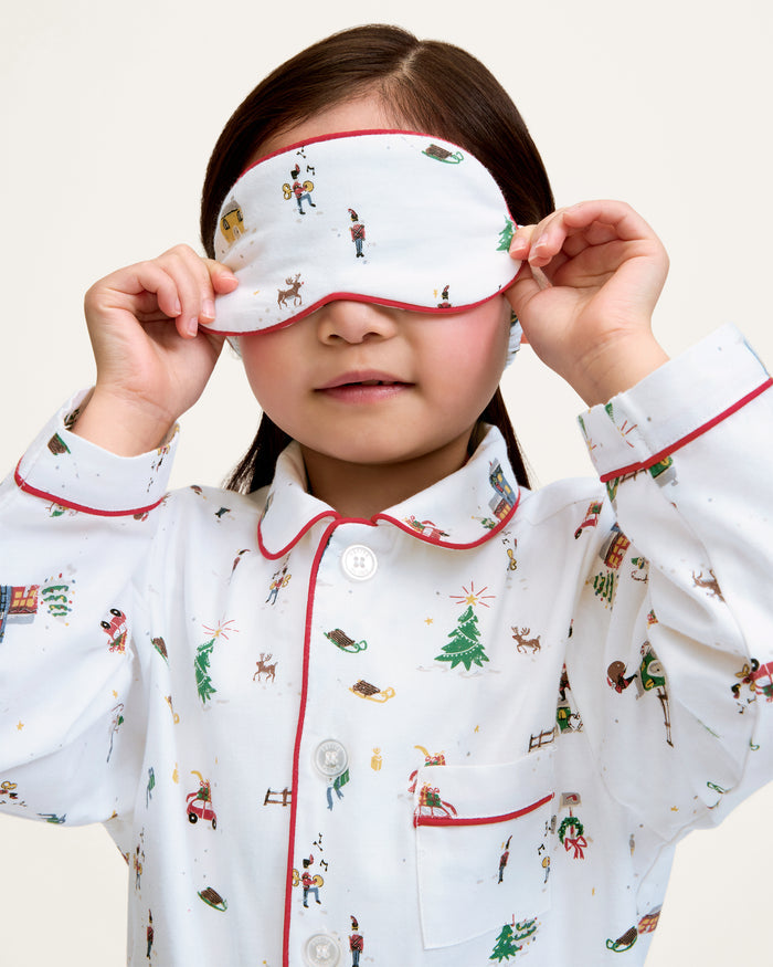 The Kid's Twill Sleep Mask in Holiday Town is a white mask with red trim, decorated with winter scenes of people, a reindeer, a house, a sled, and snowy trees. A small white tag is attached on the right side.