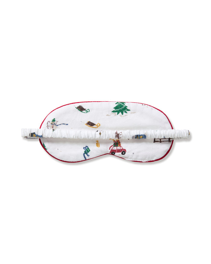 The Kid's Twill Sleep Mask in Holiday Town is a white mask with red trim, decorated with winter scenes of people, a reindeer, a house, a sled, and snowy trees. A small white tag is attached on the right side.