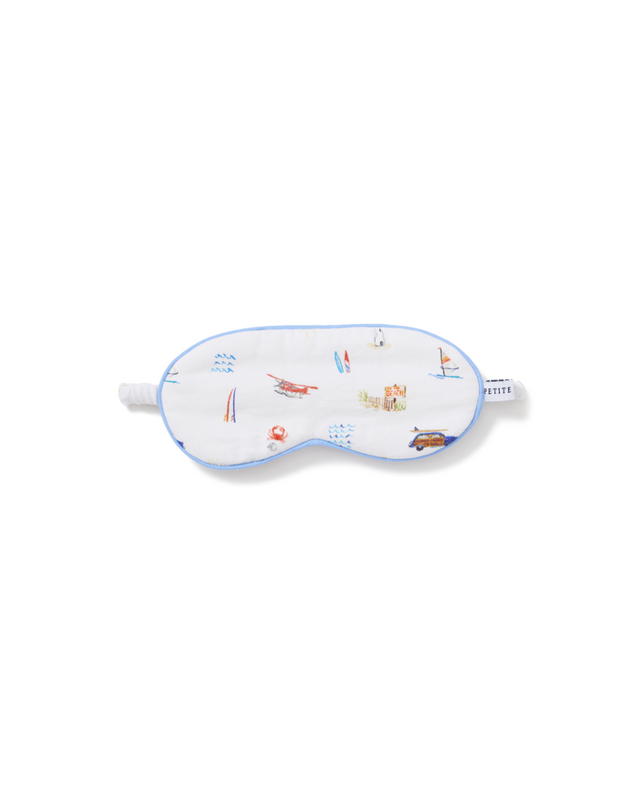 Kid’s Twill Sleep Mask in Summer Nostalgia—white with blue trim and decorated with small illustrations of beach scenes like a lifeguard stand, surfboards, boats, and a car.