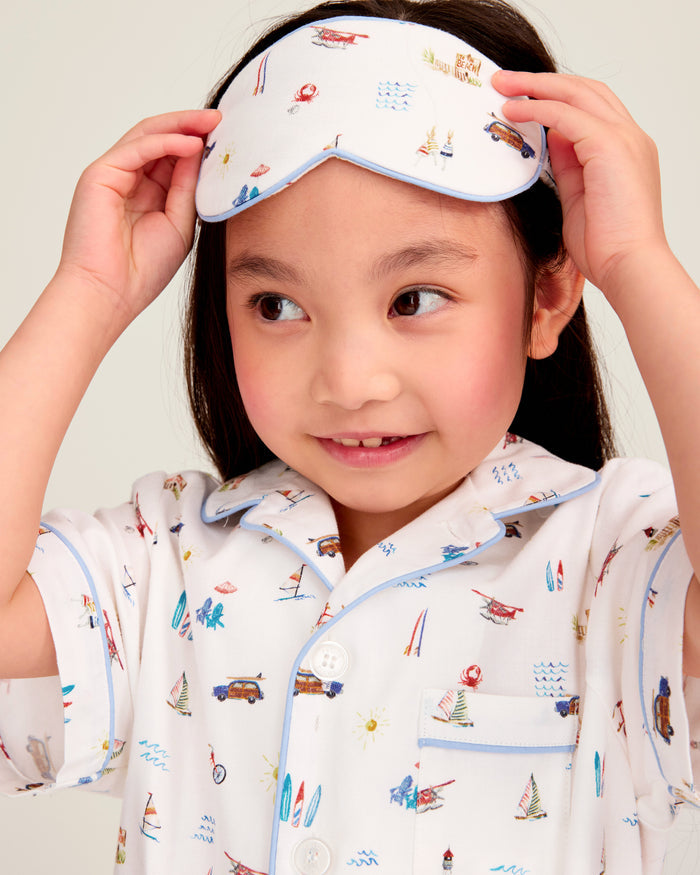 A young child in white, patterned pajamas lifts the Kid’s Twill Sleep Mask in Summer Nostalgia onto their head, smiling and looking off to the side. The pajamas feature colorful travel-themed illustrations.