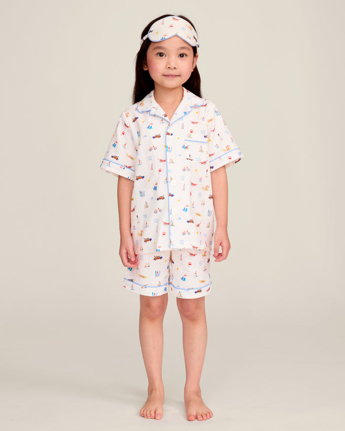 A young child stands barefoot in light-colored pajamas with a playful print, matching shorts, and the Kid’s Twill Sleep Mask in Summer Nostalgia on their head. The background is plain and neutral.