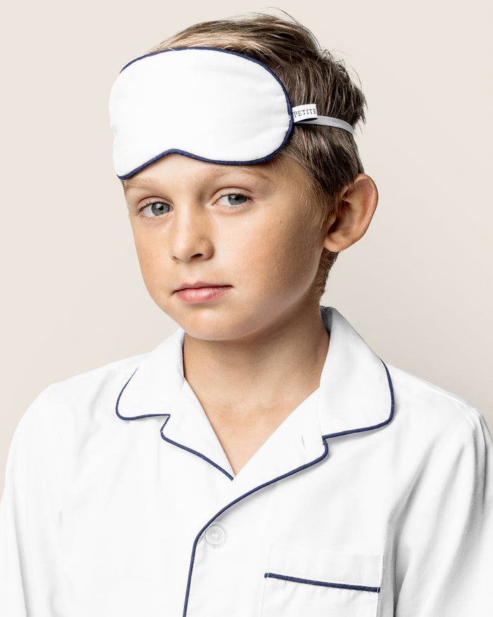 A child, in Petite Plumes Kids Twill Sleep Mask in White with Navy Piping, stands against a plain background. Their short hair complements the relaxed expression that radiates comfort and calm.