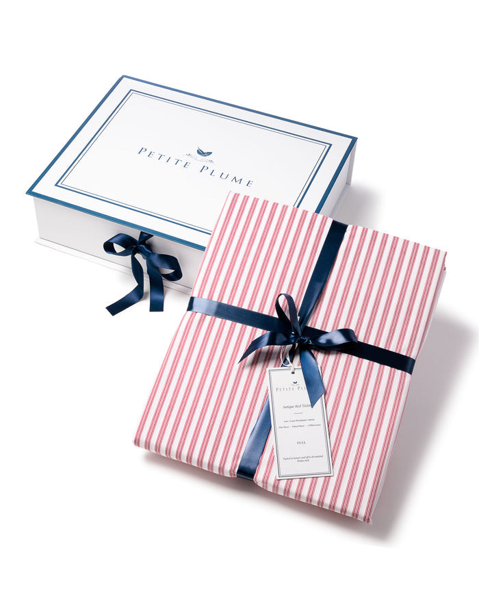 A folded Luxe Premium Cotton Bed Sheet in Antique Red Ticking by Petite Plume, resembling luxury bedding, is tied with a dark blue ribbon in front of an open white box. The box lid features a blue border and text. A tag dangles from the ribbon on this exquisite 100% cotton piece.