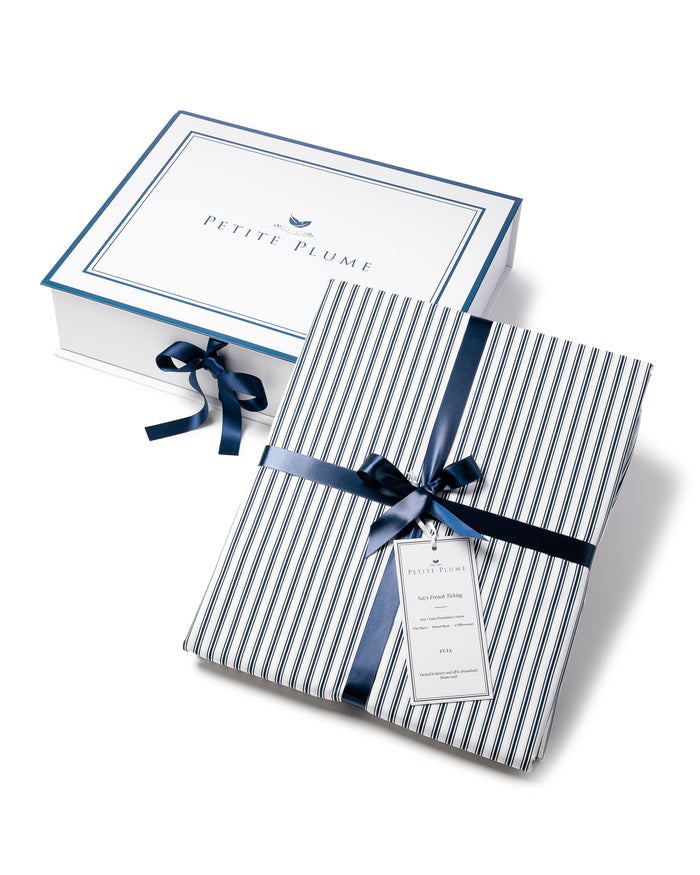 A neatly wrapped package features a navy blue and white striped pattern with a dark blue ribbon and tag, elegantly placed in front of a white box bordered in blue with PETITE PLUME at the center, echoing the luxe feel of Petite Plumes Luxe Premium Cotton Bed Sheets in Navy French Ticking.