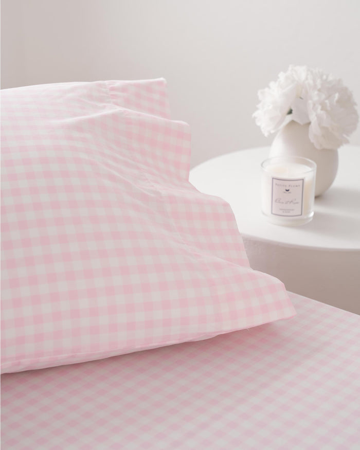 A close-up of a bed with Luxe Premium Cotton Pink Gingham Bed Sheets by Petite Plume. A round white side table with a small vase of white flowers and a scented candle completes the bright, minimalistic setting.