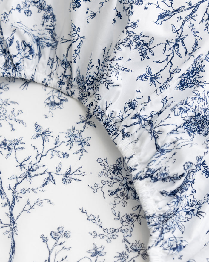 Close-up of Luxe Premium Cotton Bed Sheets in Timeless Toile, by Petite Plume, featuring intricate blue floral and foliage designs on white. The gathered fabric highlights vintage-inspired elegance, ideal for luxury bedding enthusiasts.