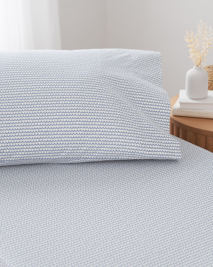The bed is adorned with Petite Plumes Luxe Premium Cotton La Mer sheets, featuring a white and blue pattern. Nearby, a small wooden side table holds a white vase with dried flowers and stacked books as soft light filters through the window.