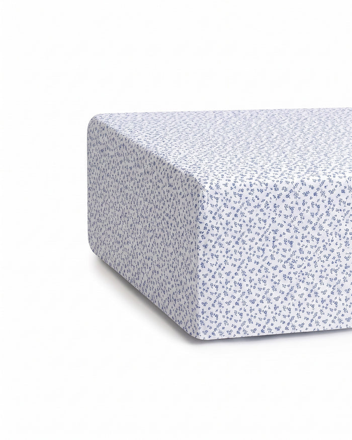 A close-up of a mattress corner covered with the Luxe Premium Percale Crib Sheet in Ditsy Waltz White, featuring a small blue floral pattern. The sheet fits snugly around the mattress.