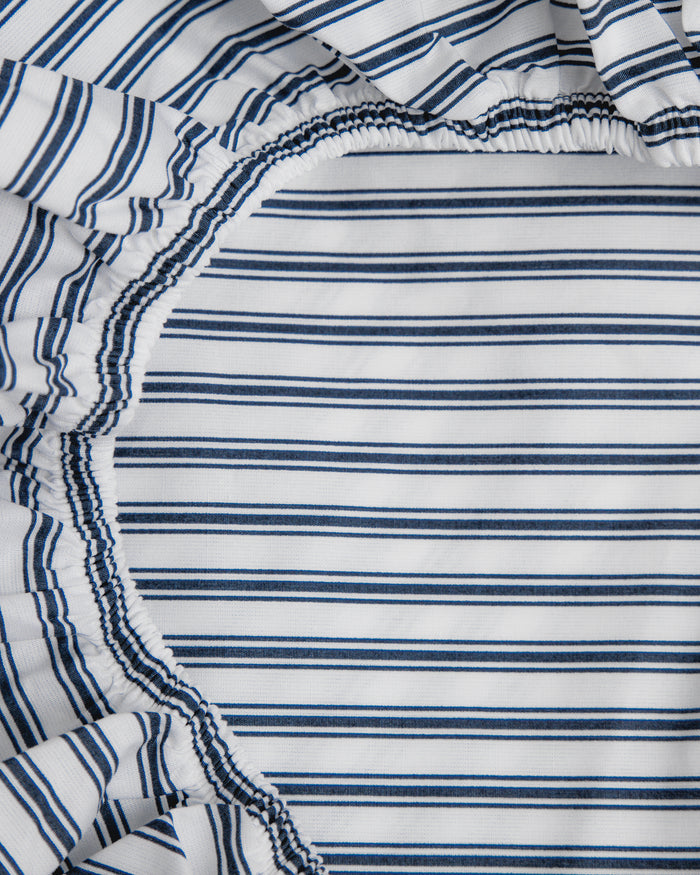 A close-up of Petite Plumes Luxe Premium Cotton Crib Sheet in Navy French Ticking shows breathable cotton percale with thin blue and white stripes, gathered with elastic into a circular shape at the corner.