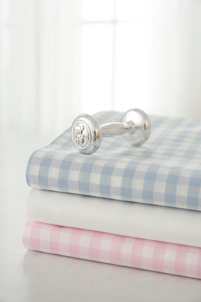 A silver baby rattle sits on three folded fabrics, one being the Luxe Premium Cotton Pink Gingham Crib Sheet by Petite Plume, all stacked on a table. A softly lit window with sheer curtains in the background enhances the blue, white, and pink checkered pattern, creating a bright and serene atmosphere.