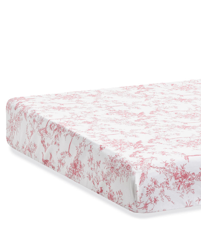 A close-up of the Luxe Premium Percale Crib Sheet in Pink Timeless Toile, featuring a red and white floral toile pattern. The fitted sheet hugs the mattress snugly, with corners neatly tucked in.