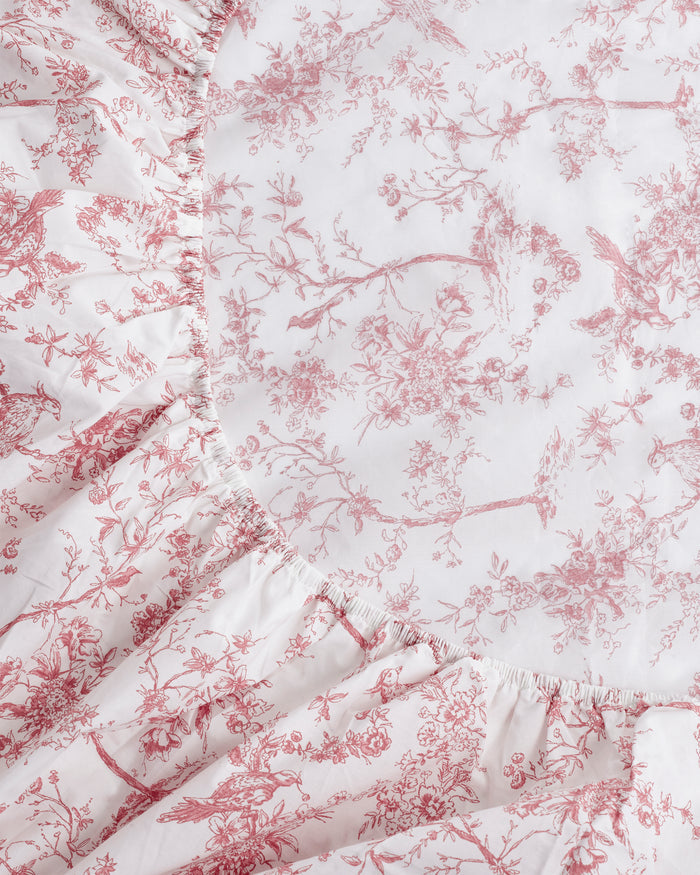 A close-up of the Luxe Premium Percale Crib Sheet in Pink Timeless Toile, featuring a red and white floral toile pattern. The fitted sheet hugs the mattress snugly, with corners neatly tucked in.