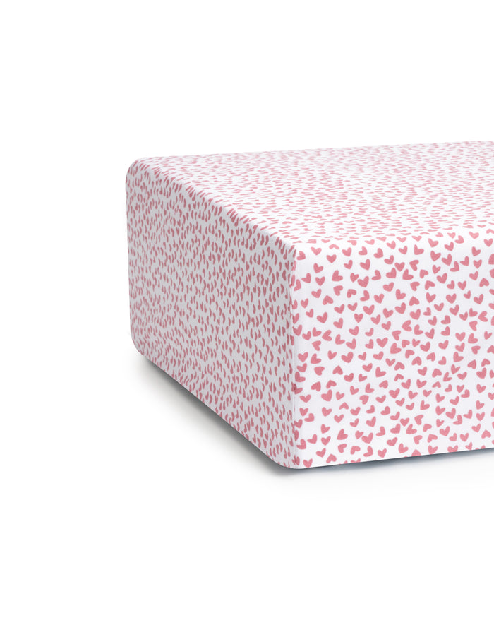 The Luxe Premium Sweethearts Crib Sheet by Petite Plume features ultra-soft, white fabric adorned with small pink heart patterns, evenly distributed for a charming and playful design. The image highlights the corner, showcasing the delightful pattern like cotton percale furniture.