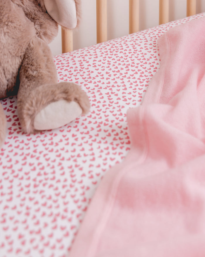 The Luxe Premium Sweethearts Crib Sheet by Petite Plume cradles a soft brown stuffed animal on an ultra-soft mattress, its breathable cotton percale featuring small pink hearts. A cozy pink blanket drapes over the side, adding a gentle touch.