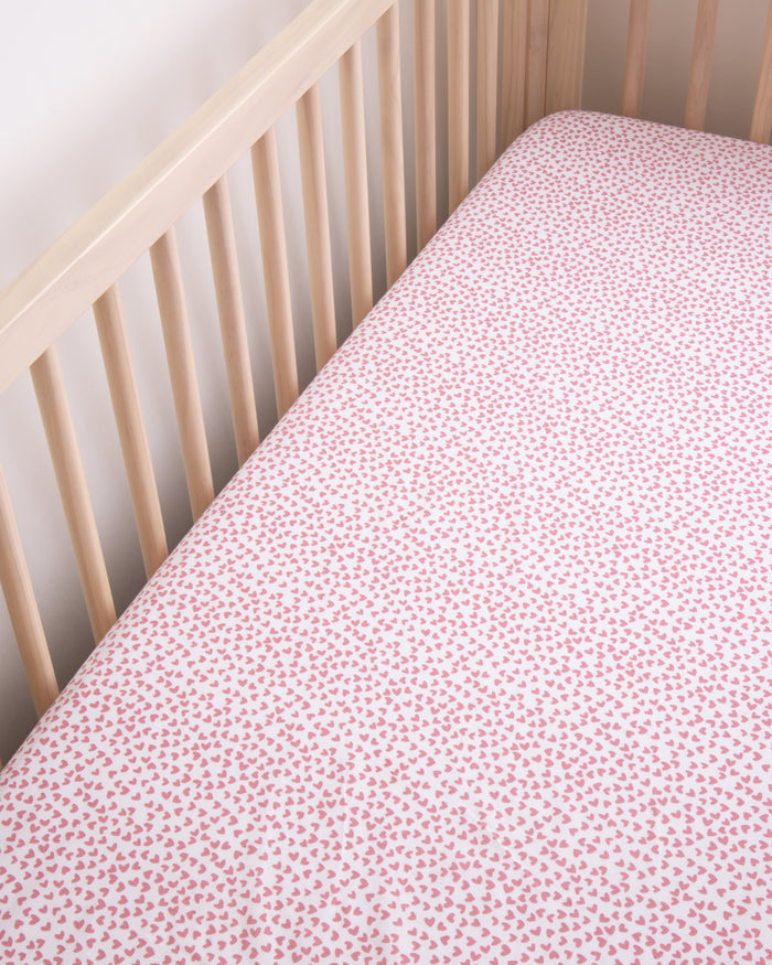 The Luxe Premium Sweethearts Crib Sheet by Petite Plume is an ultra-soft, breathable cotton percale in a pink and white heart pattern. Paired with a wooden crib against a neutral wall, it perfects the soft and cozy nursery setting.