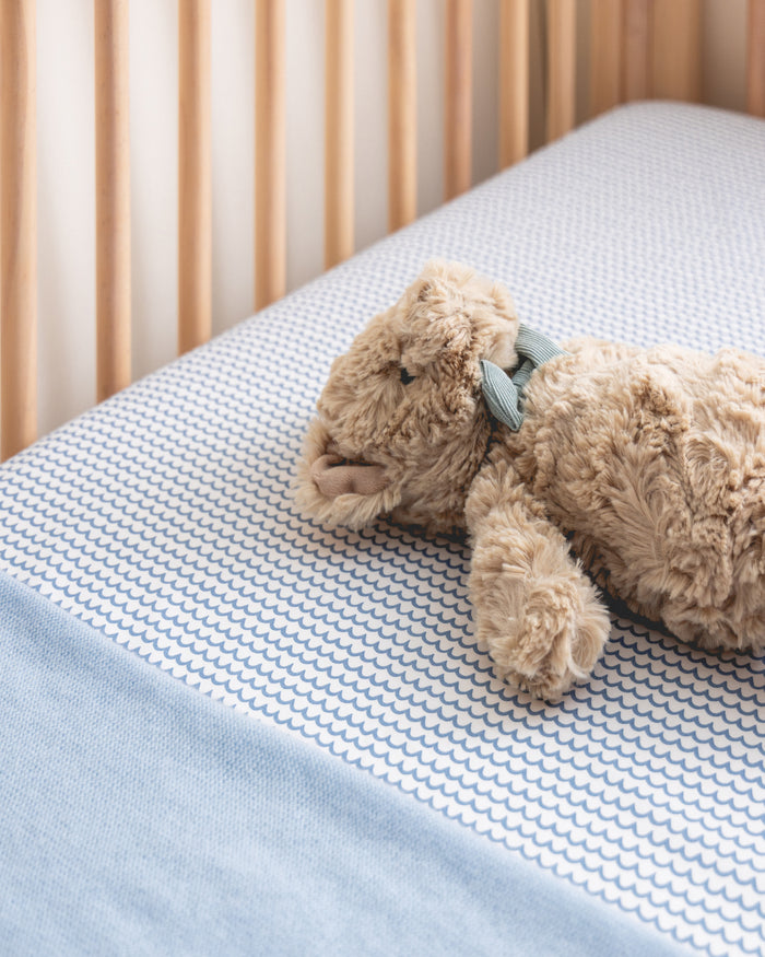 A plush teddy bear with a blue bow rests on a crib mattress topped with the Luxe Premium La Mer Crib Sheet by Petite Plume, paired with a matching blue blanket. This setup creates a soft and cozy nursery atmosphere.