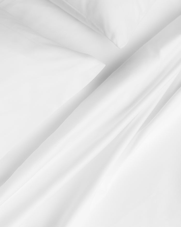 A close-up of a neatly made bed showcases the Luxe Premium Percale Duvet Set in White by Petite Plume. The 100% cotton fabric is smooth and crisp, exuding cleanliness and comfort. The image highlights where the pillow meets the folded percale duvet.