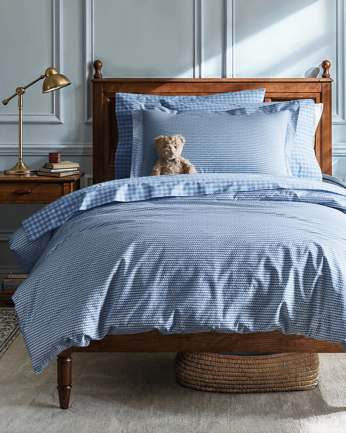 A cozy bedroom scene features a wooden bed adorned with the Luxe Premium Percale Duvet Set in La Mer by Petite Plume. A plush teddy bear leans on the pillows. Beside it, a wooden nightstand holds a brass lamp, books, and a clock as soft natural light streams through the window.