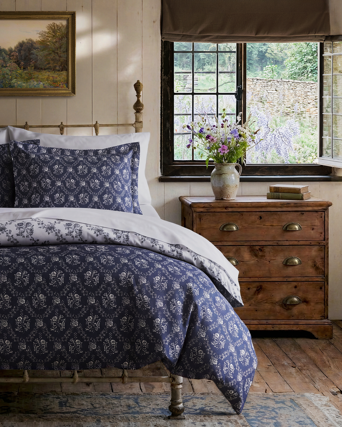 A cozy bedroom featuring a metal bed frame, the Luxe Premium Percale Reversible Duvet Set in Innkeeper Trellis, a wooden nightstand, vase of flowers, rustic window, and a framed landscape painting on a wood-paneled wall.