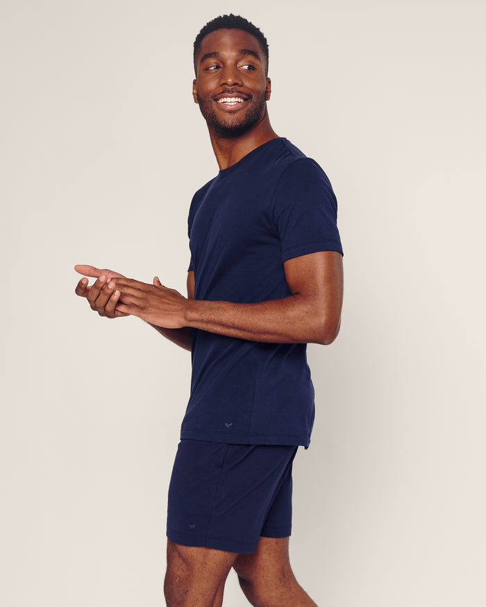 A man smiles, looking right while wearing Petite Plumes Mens 5 Pima Shorts in Navy and a matching t-shirt, made from soft Peruvian Pima cotton. He stands against a light-colored background with his hands clasped in front.