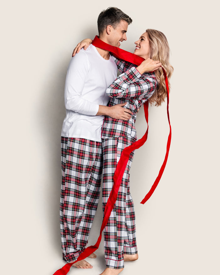 A joyful couple in matching Petite Plumes Balmoral Tartan pajamas wraps a long red ribbon around themselves. The man sports Mens Brushed Cotton Pants with a white long-sleeve shirt, while the woman wears a matching pajama top. They stand close, smiling at each other.