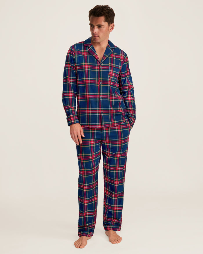 A man stands barefoot wearing the Men’s Brushed Twill Pajama Set in Windsor Tartan, featuring a matching long-sleeve, button-up top and pants with a blue, red, and green plaid pattern. He looks slightly to the side against a plain background.