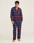 A man stands barefoot wearing the Men’s Brushed Twill Pajama Set in Windsor Tartan, featuring a matching long-sleeve, button-up top and pants with a blue, red, and green plaid pattern. He looks slightly to the side against a plain background.