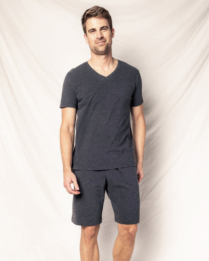 In front of a light fabric backdrop, a person wearing Petite Plumes Mens Classic Pima Shorts in Dark Heather Grey and a matching V-neck T-shirt smiles with their arms relaxed by their side, showcasing comfort and style with the softness of Peruvian Pima cotton.