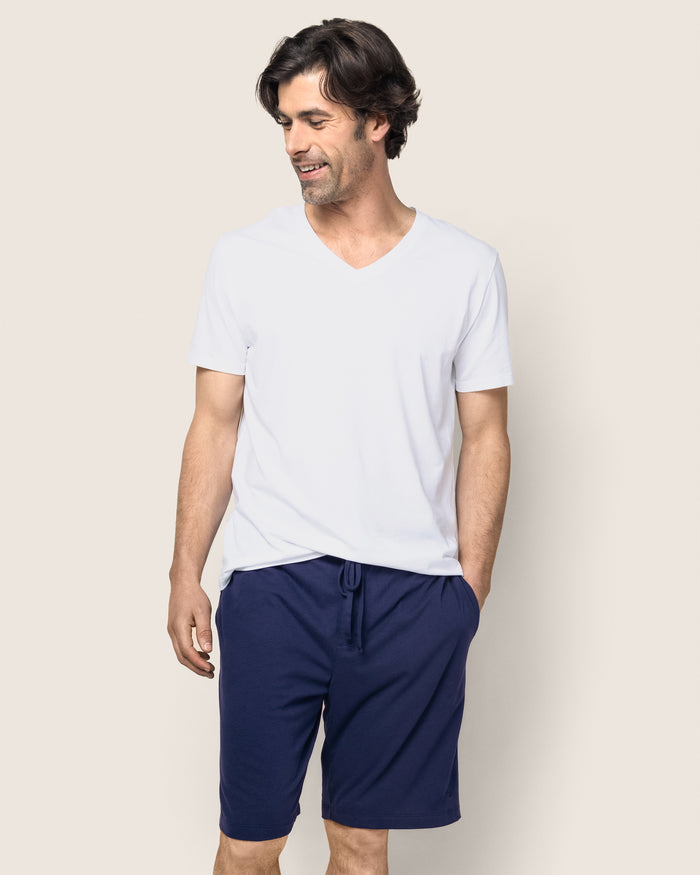 A man with dark hair, wearing a white V-neck T-shirt and Petite Plumes Mens Classic Pima Shorts in Navy, smiles against a plain light background, one hand in his pocket, enjoying the comfort of his bedtime ritual attire.