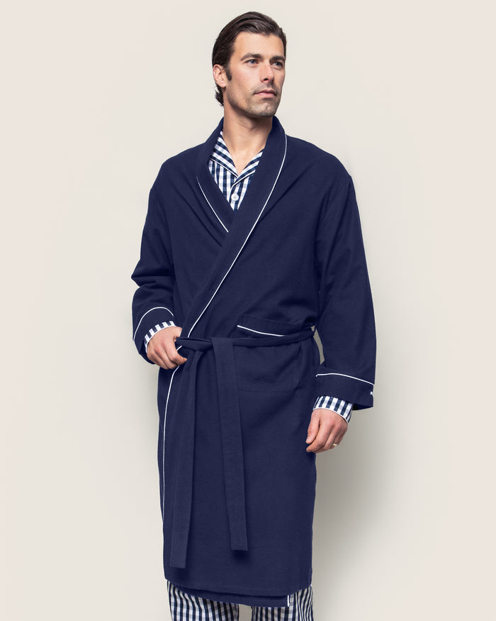 A man wears Petite Plumes Mens Flannel Robe in Navy with White Piping, featuring moisture-wicking fabric and a tie belt over striped pajamas. He stands against a plain background, gazing to the side with a calm expression.