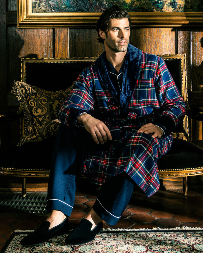 A man sits on a luxurious sofa wearing a red and blue plaid robe over dark mens pajamas and Petite Plumes Mens Hamptons Loafer in Black. The setting is opulent, featuring wooden paneling and a decorative cushion, creating a sophisticated atmosphere.
