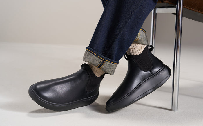 A person wearing black Chelsea-style ankle boots with dark blue jeans and striped socks, seated near a metal chair leg on a light studio floor | lifestyle