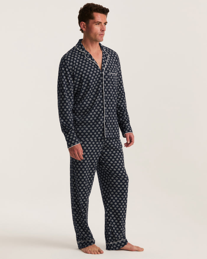 A man stands barefoot wearing the Men’s Lightweight Pima Pajama Set in Captain’s Compass, featuring dark blue pajamas with a white geometric pattern. The pajama top includes a collar, long sleeves, buttons, and a chest pocket. The background is plain and light-colored.